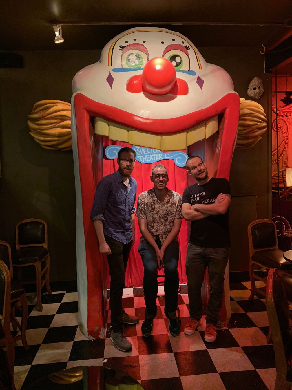 Scott Simpson, Ben Jennings, and Jon Allen standing inside a bizarre clown in a dimly lit theater lobby.