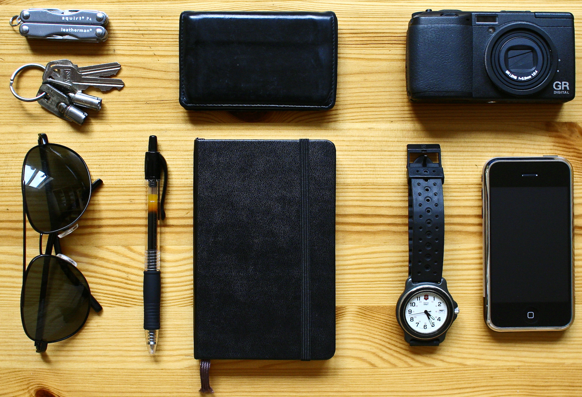 A top-down view of the following items, arranged neatly on a wooden tabletop: a small Leatherman pocketknife/multi-tool, a keyring with four keys, a small leather wallet, a Ricoh GR-D digital camera, cheap black aviator sunglasses, a 0.5 mm Pilot G2 pen, a black pocket-sized hardcover Moleskine notebook, a black Swiss Army wristwatch, and an original iPhone.