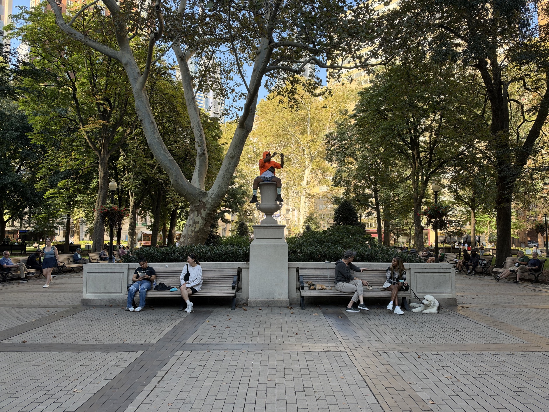 Afternoon in Rittenhouse Park, Philadelphia, on lovely late summer day. A man (himself taking photos or photo) sits high atop a planter between park benches.